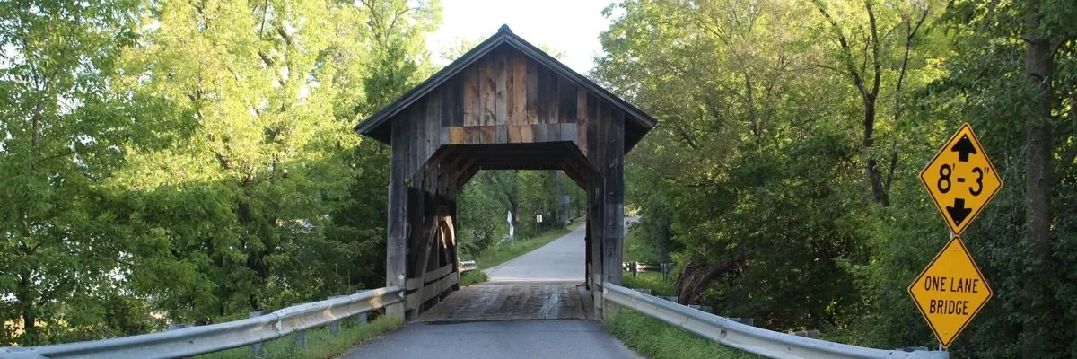A wooden covered bridge with a road passing through it, surrounded by trees and a 'One Lane Bridge' sign.