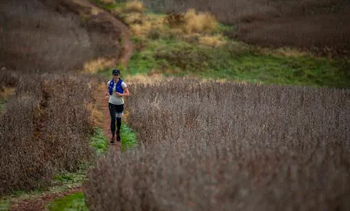 A solo runner on a winding dirt trail through dry brush and green hills.