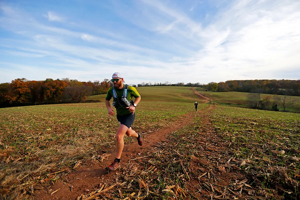 A runner with a hydration pack runs on a dirt trail through a field with rolling hills and trees in the background under a partly cloudy sky.