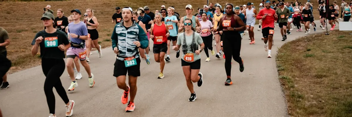 A group of runners in a race, with bib numbers visible, running on a paved path with grassy areas on the sides.