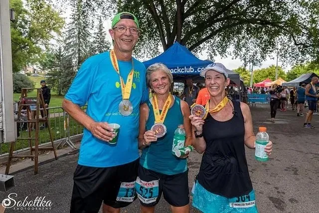 Three smiling runners, two women and one man, pose for a photo after a race, all wearing medals and holding drinks.