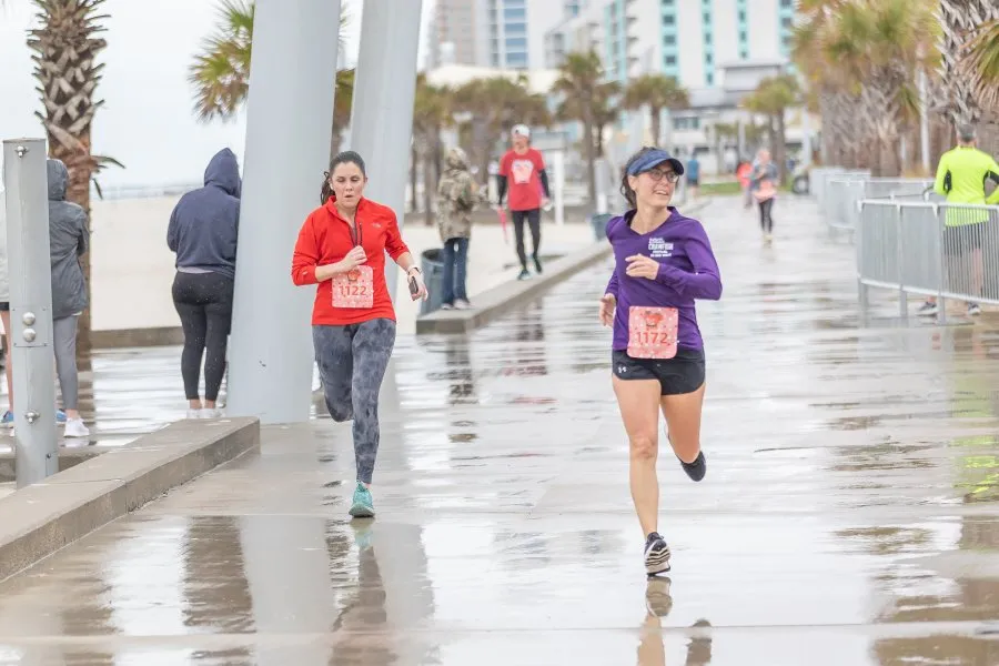 Two women running on a wet, paved path next to a beach with palm trees and buildings in the background.