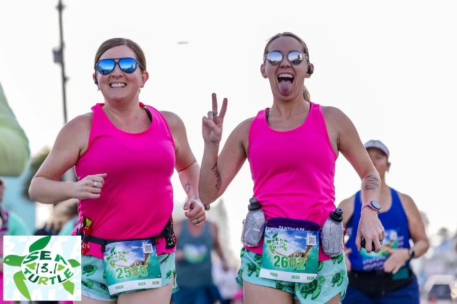 Two female runners in bright pink tank tops smile and pose for the camera during a race, one giving a peace sign and sticking her tongue out.