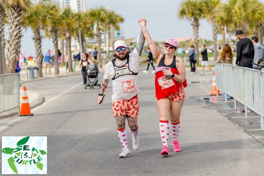 A male and female runner cross the finish line with their arms raised in celebration, holding hands.