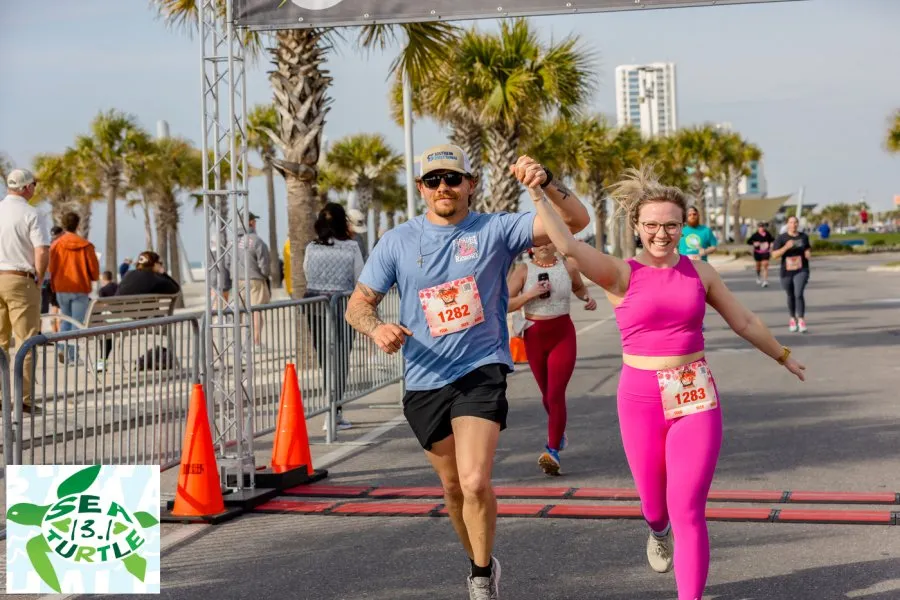 A male and female runner, holding hands and smiling, cross the finish line of a road race. The male runner wears a blue t-shirt and sunglasses, while the female runner wears a bright pink outfit. Palm trees and a coastal city skyline are visible in the background.