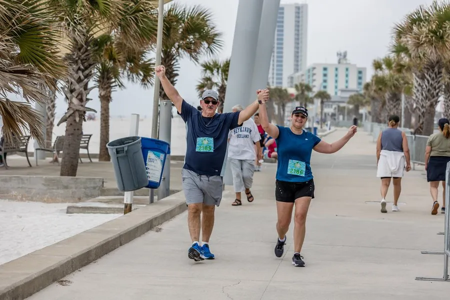 A man and a woman, both runners, cross the finish line holding hands and raising their arms in celebration, with palm trees and buildings in the background.