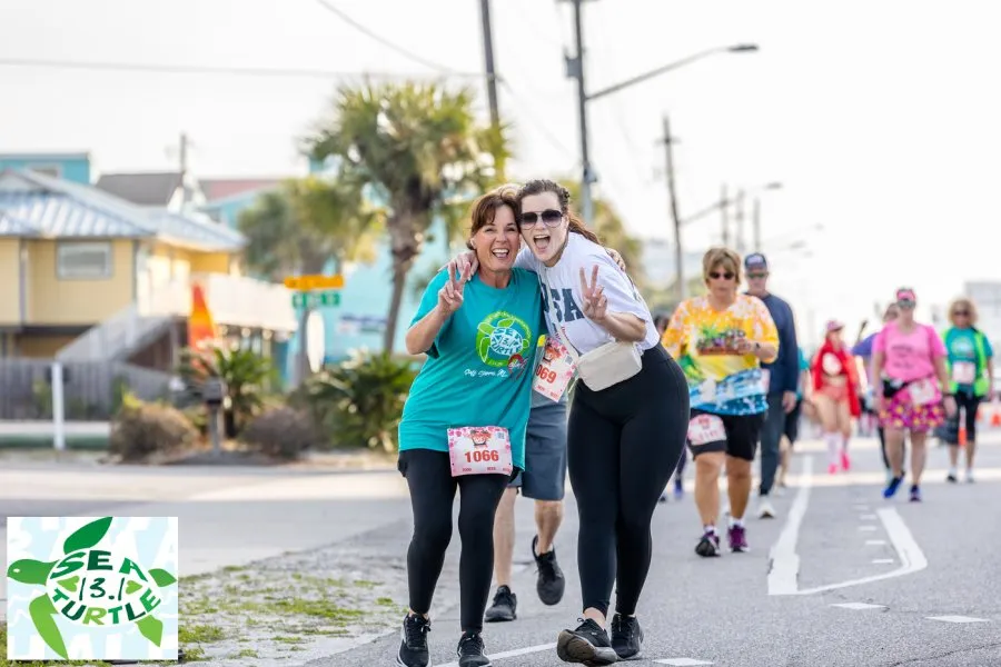 Two women smiling and giving peace signs while running in a race, with other participants in the background.