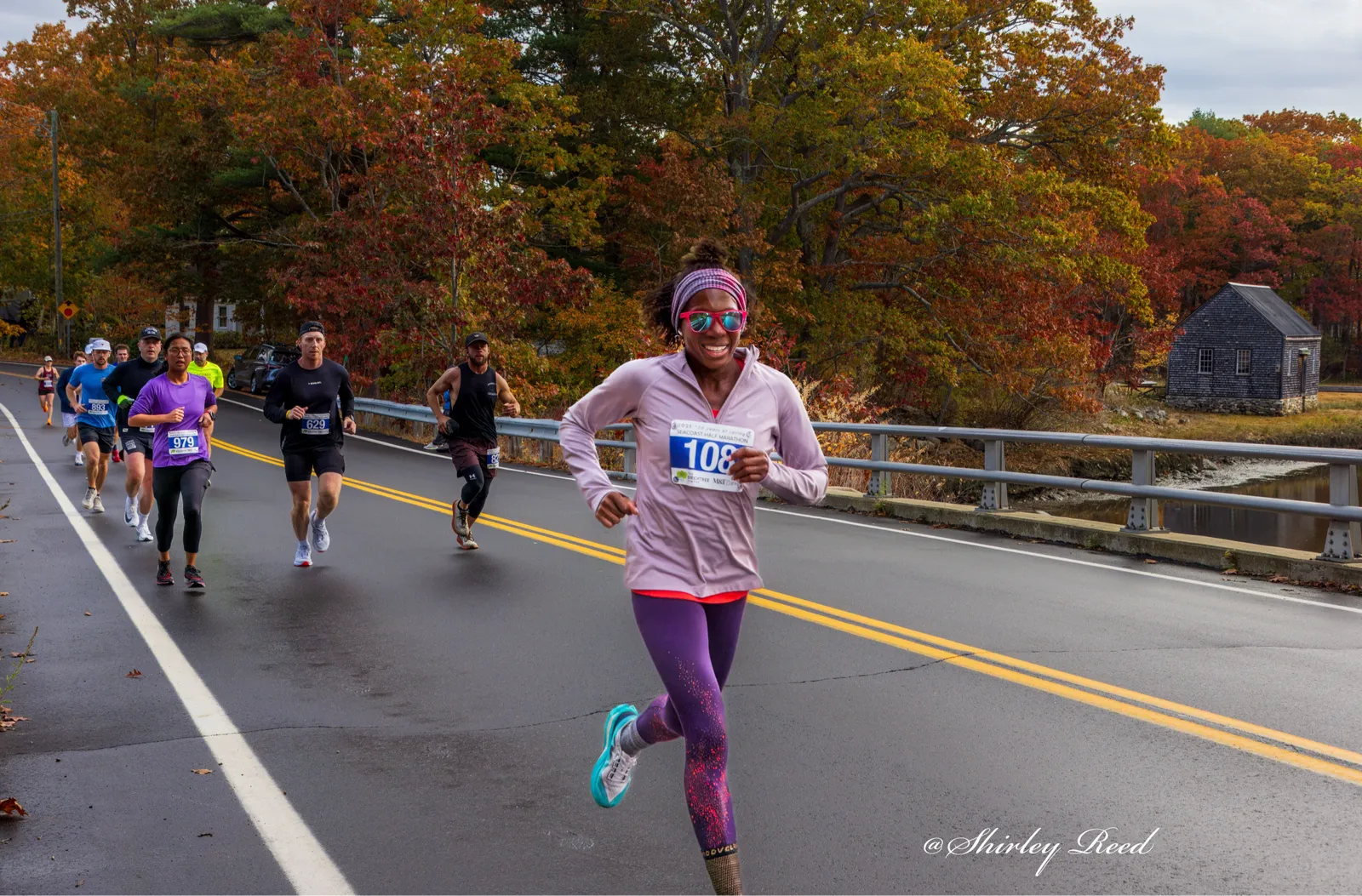 A female runner smiles as she runs on a road with autumn foliage in the background.