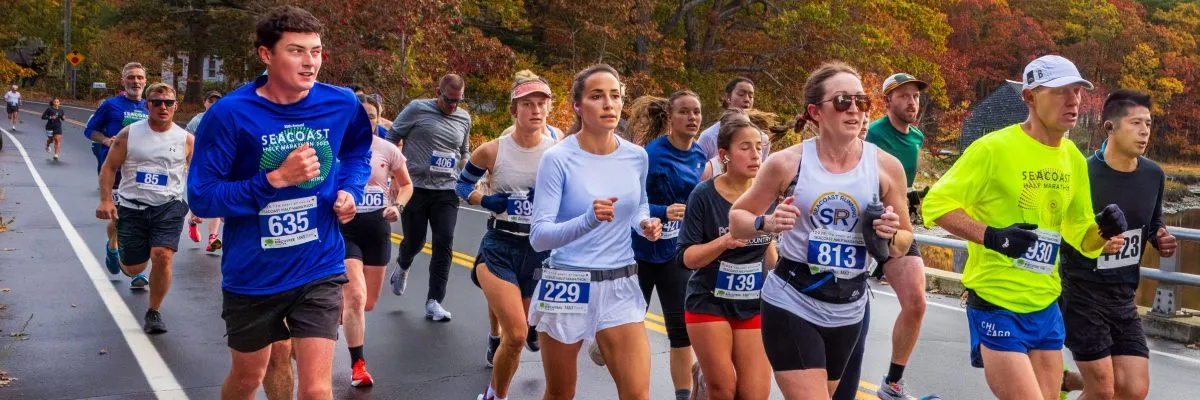 Runners participating in the Seacoast Half Marathon, with some wearing race bibs and branded shirts, running on a road lined with autumn trees.