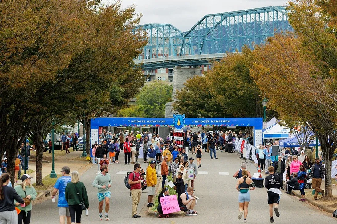 Runners gather at the start line under the '7 Bridges Marathon' arch, with a large blue bridge in the background.