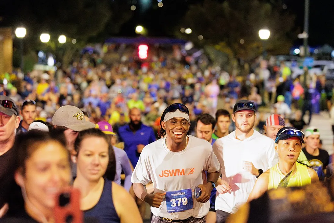A diverse group of runners, with a smiling man in the foreground wearing a t-shirt with bib number 379, running at night during the Seven Bridges Marathon.