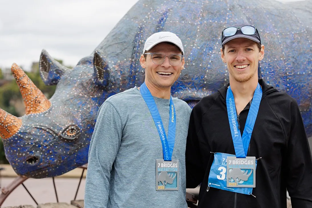 Two smiling male runners wearing medals from the Seven Bridges Marathon stand in front of a large, blue, mosaic rhinoceros sculpture.