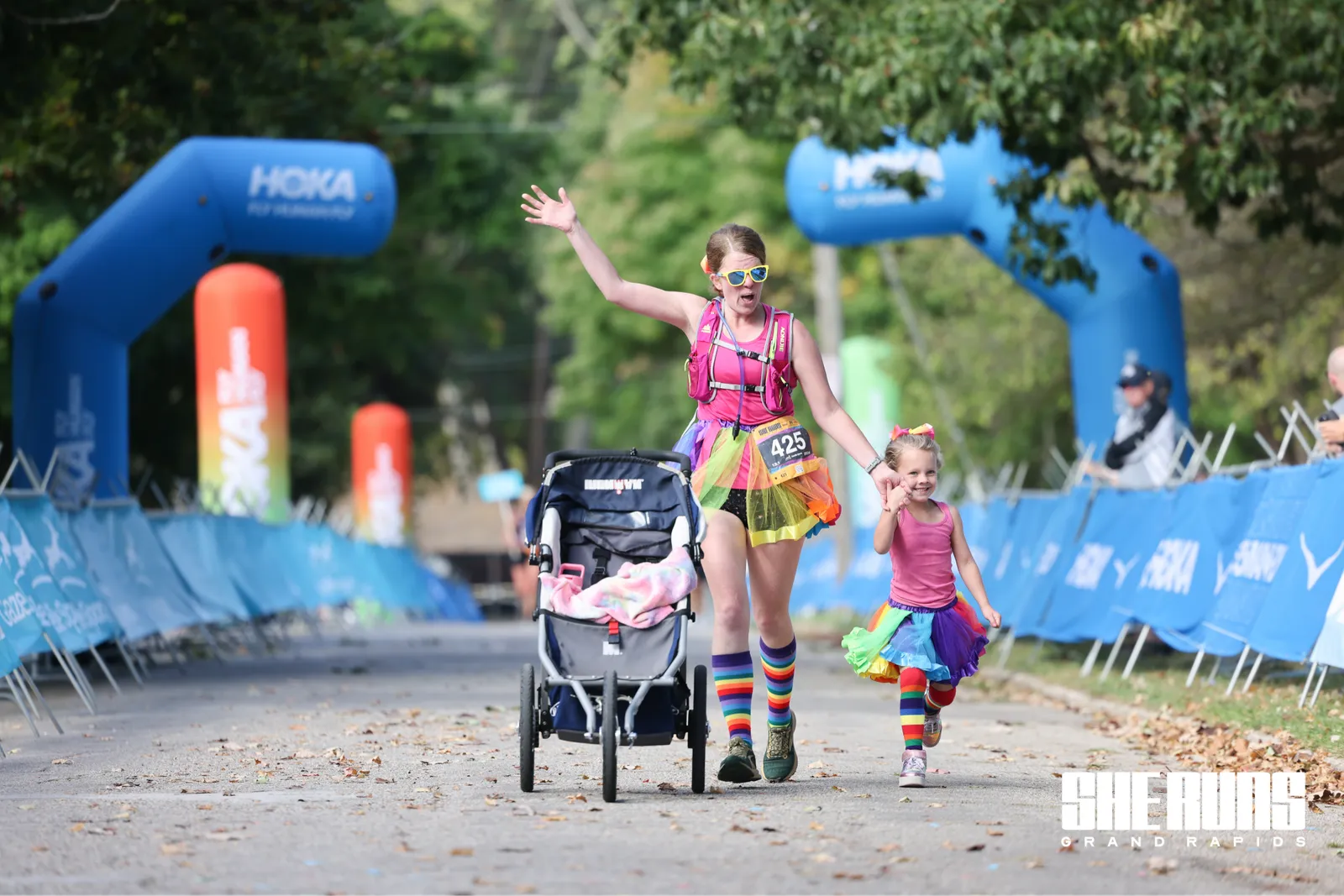 A woman and a young girl run down a road, both wearing colorful outfits and rainbow socks. The woman is pushing a stroller and waving, while the girl holds her hand and smiles.