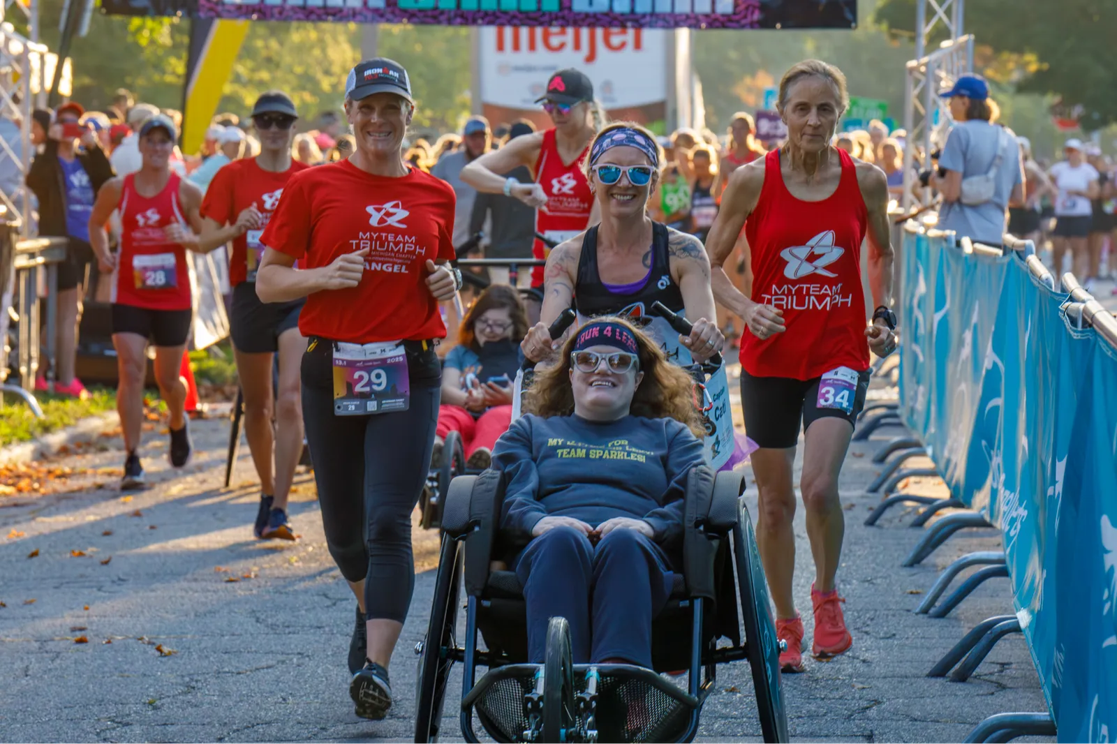 A group of runners, including a participant in a wheelchair, smiling as they run together at the start of a race.