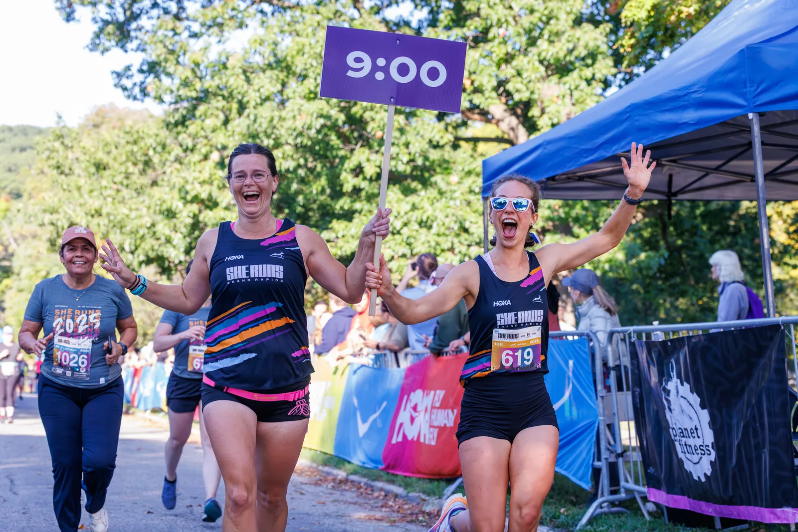 Two women running and cheering at a race, one holding a sign that says '9:00'.