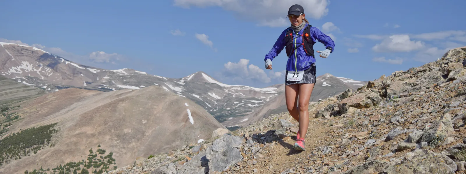 A female runner with bib number 15 descends a rocky mountain trail under a blue sky with scattered clouds.
