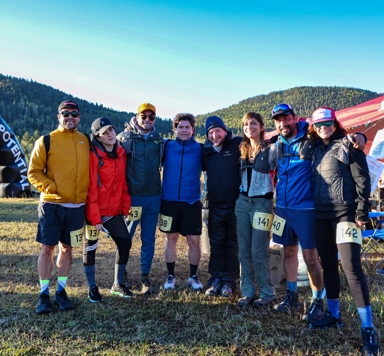 A group of eight runners pose for a photo at the start line of a trail race, with mountains in the background.