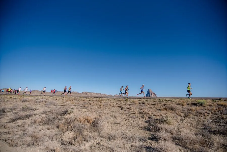 Runners race along a road with a distinctive rock formation in the background under a clear blue sky.
