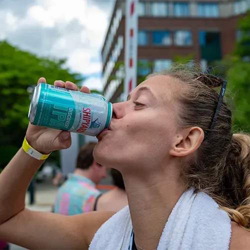A woman wearing a yellow race bib drinks from a can of Shipyard IPA after a race.