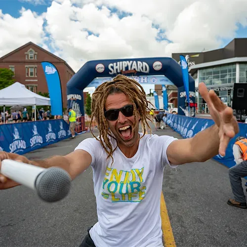 An excited man with dreadlocks holds a microphone and gestures towards the camera in front of a Shipyard finish line arch.