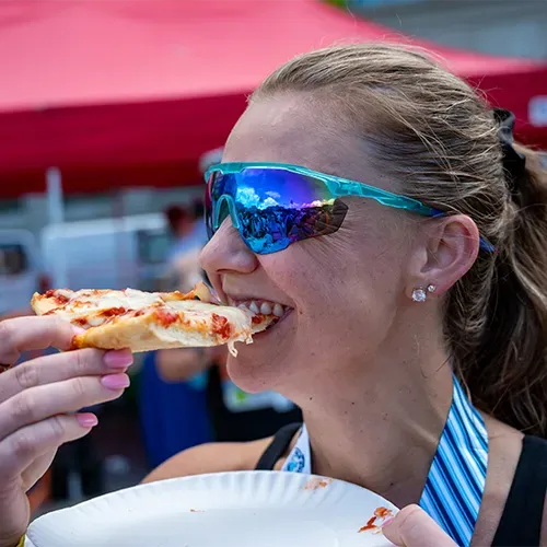 A woman wearing sunglasses and a race bib eats a slice of pizza after a race.