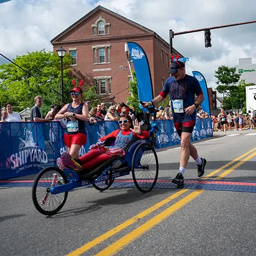 A runner pushes a participant in a racing wheelchair across the finish line, both smiling and waving.