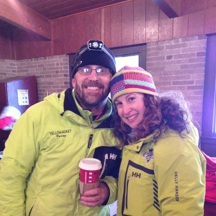 A man and a woman smiling at the camera, both wearing bright yellow jackets and winter hats. The man is holding a Starbucks cup.