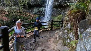 Two runners on a wooded trail with a waterfall in the background.