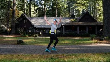 A female runner with her arms raised in celebration runs on a paved path in front of a rustic lodge surrounded by trees.