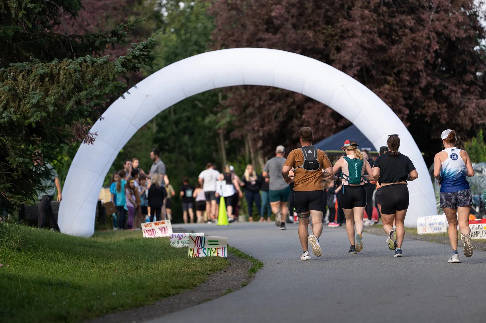 Runners approaching a white inflatable archway on a paved path, with spectators and signs visible in the background.