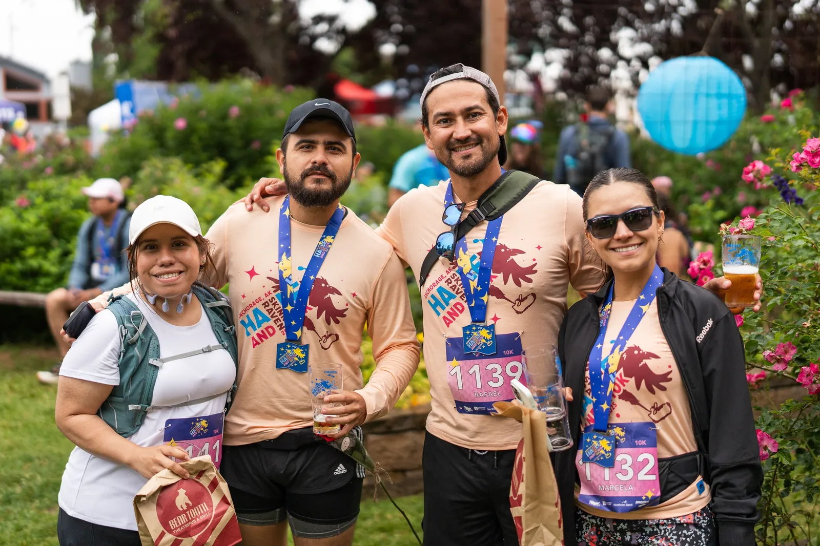 Four smiling runners, two men and two women, pose for a photo after a race, wearing medals and holding drinks.