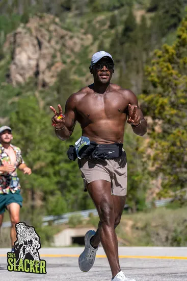 A shirtless runner with a determined expression runs on a road, holding up peace signs with both hands, with scenic mountains and trees in the background.