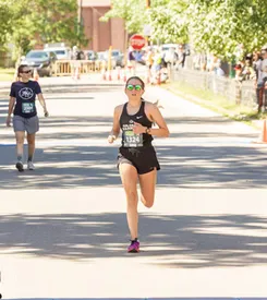 A female runner with sunglasses and bib number 1324 runs towards the camera on a paved road, with another runner and spectators in the background.