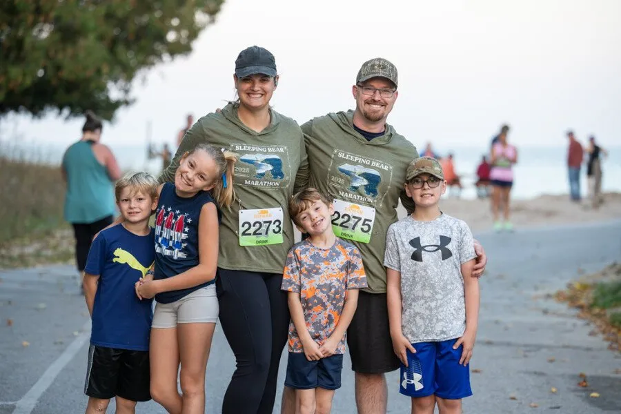 A smiling family of five, including two parents and three children, pose for a photo at a race. The parents are wearing race t-shirts with bib numbers, and the children are dressed in athletic wear. Other runners and spectators are visible in the background.