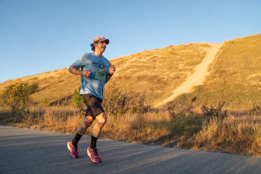 A male runner with bib number 104 runs on a paved road with a grassy hill in the background under a clear blue sky.