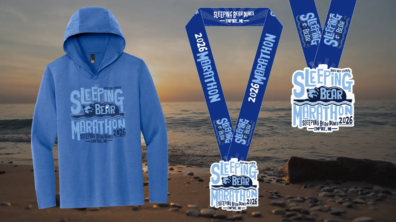 Sleeping Bear Marathon 2026 merchandise including a hooded shirt and medal, displayed on a beach with the ocean in the background.