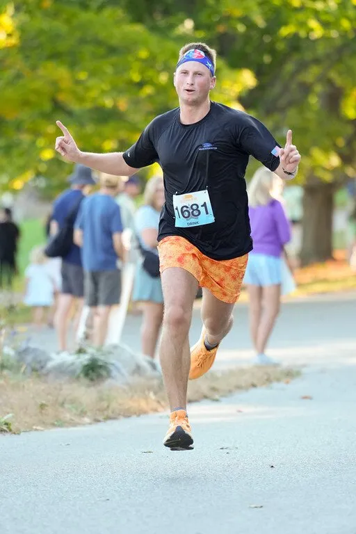 A male runner in an orange patterned shorts and black t-shirt runs towards the camera, holding up two fingers on each hand.