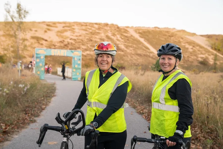 Two women in yellow safety vests and helmets stand with their bicycles in front of a race start arch.