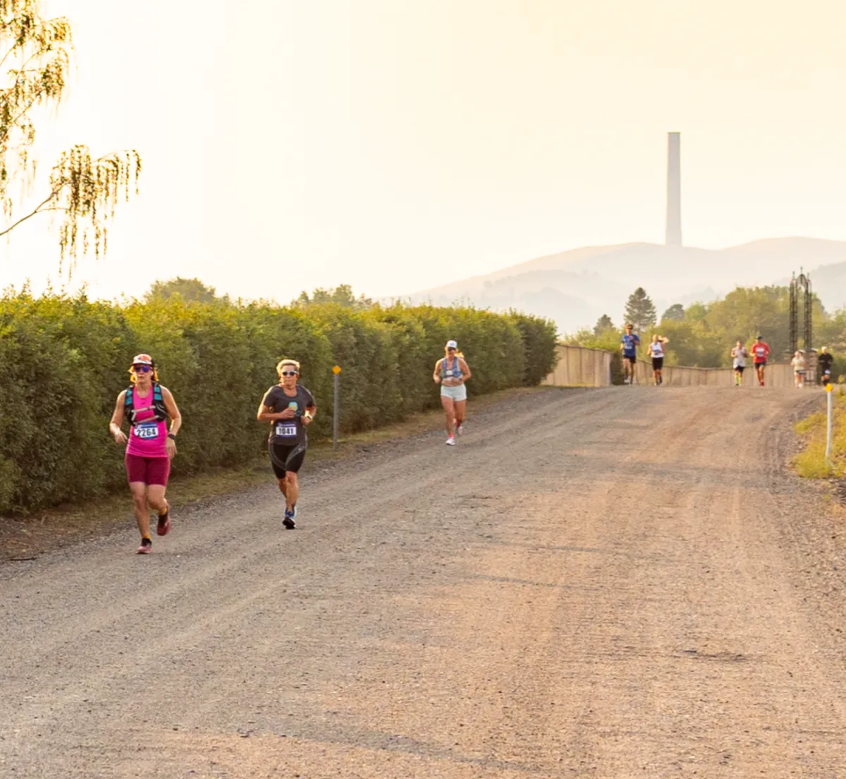 Runners on a gravel road with hills and a tall structure in the background.