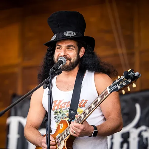 A man with long dark hair and a top hat plays a guitar and sings into a microphone at an outdoor event.