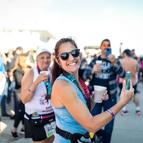 A woman smiles as she takes a selfie with her phone, wearing sunglasses and a race medal around her neck, with other runners and a crowd in the background.