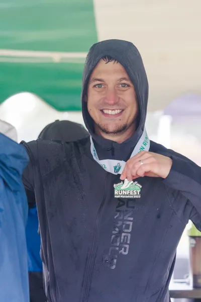 A smiling man wearing a hooded jacket and a race medal around his neck, holding the medal up to show it off.