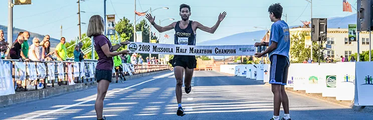 A male runner with arms raised in celebration crosses the finish line of the 2018 Missoula Half Marathon, holding a banner.