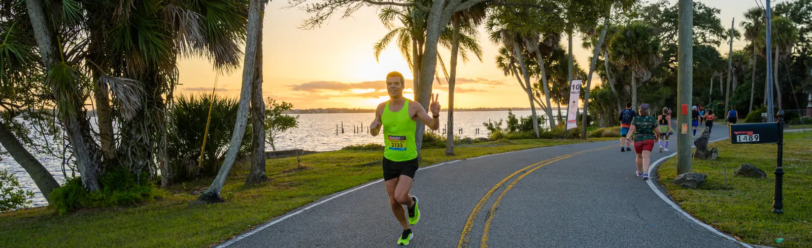 A male runner in a neon green tank top smiles and holds up two fingers while running on a road next to a body of water at sunrise.