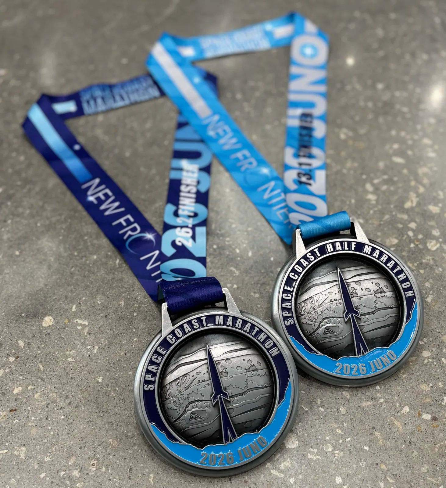 Two race medals for the Space Coast Marathon and Half Marathon, featuring rocket designs and the year 2026.