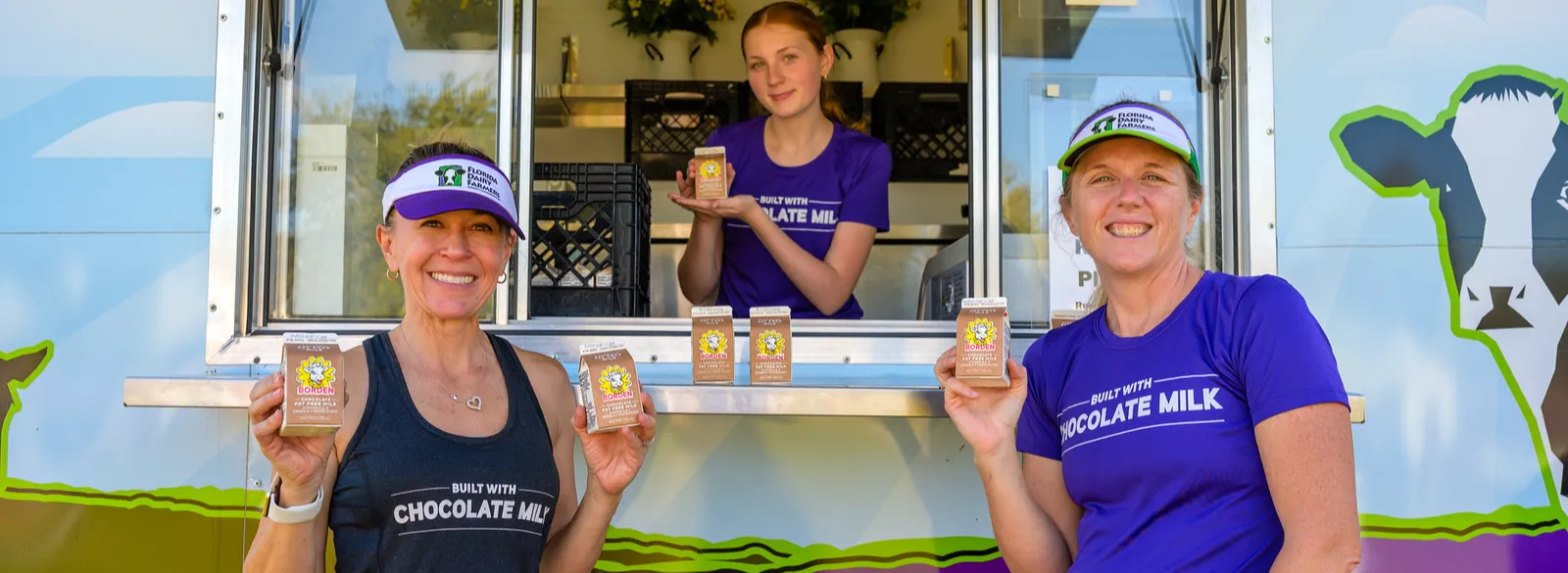Three women smiling and holding cartons of chocolate milk in front of a food truck with a cow graphic.