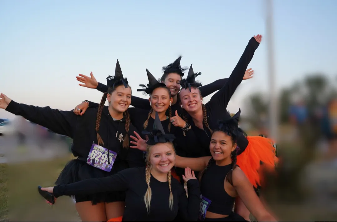 A group of six young women in black witch costumes, some wearing orange tutus, pose for a photo with smiles and arms outstretched at a race event.