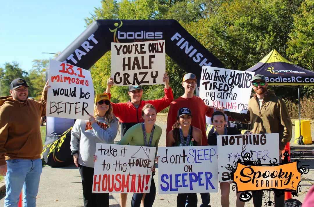 A group of smiling people stand in front of a race arch, holding up various funny signs related to running and the 'Spooky Sprint' event.
