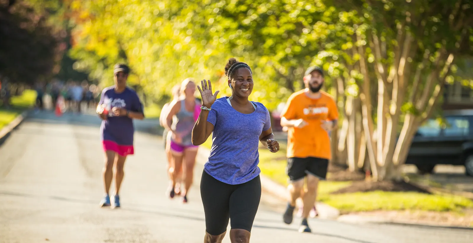 A woman smiles and waves while running in a race, with other runners behind her on a tree-lined road.