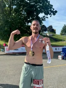 A shirtless male runner flexes his bicep and proudly displays his marathon medal after finishing the race.